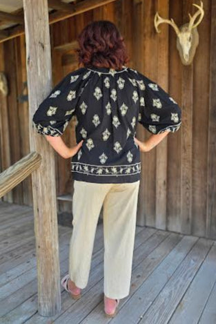 Person wearing a black patterned top and beige pants standing in a wooden interior.