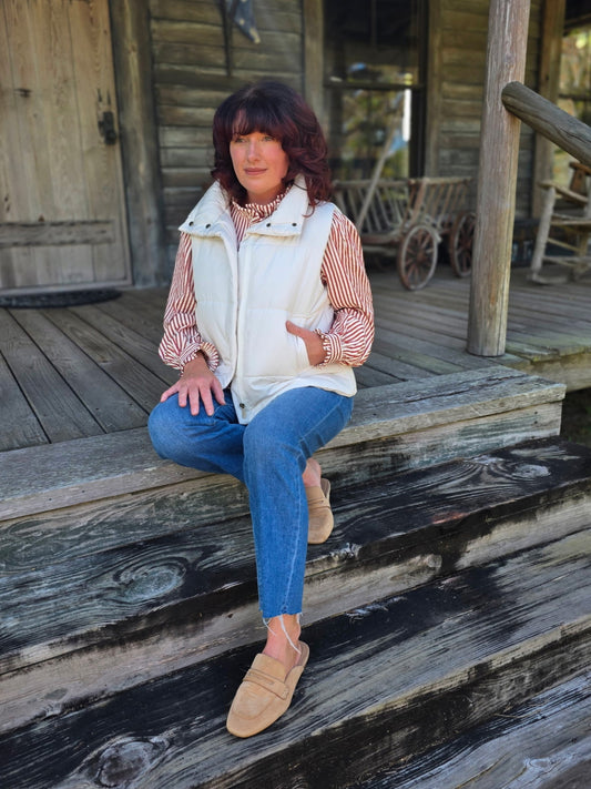 Woman wearing an ivory zip-up puffer vest styled with a striped blouse and denim jeans, standing on a rustic porch.