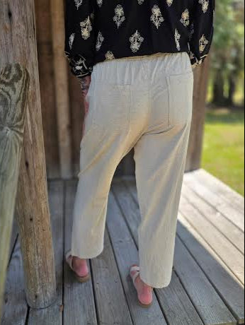 Woman wearing light beige linen pants with a drawstring waist and side pockets, styled with a black printed blouse and woven slide sandals.