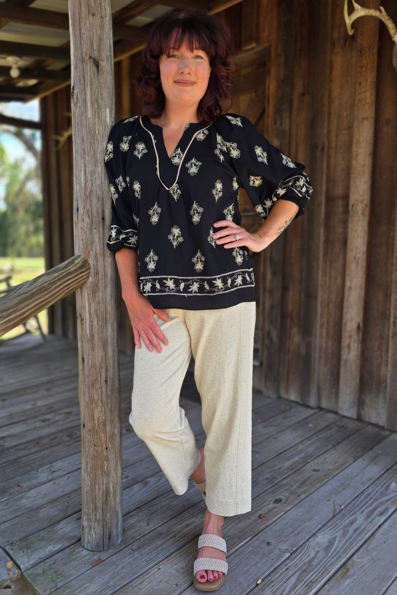 Woman standing on a wooden porch wearing a black patterned top and beige pants.