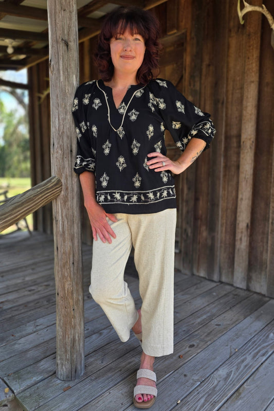 Woman standing on a wooden porch wearing a black patterned top and beige pants.