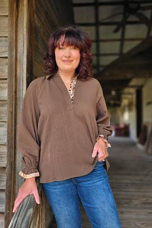 Woman in a brown shirt and blue jeans standing in front of a wooden building.