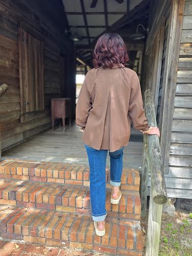 Person in a brown coat and blue jeans standing on a wooden porch with a rustic building in the background.