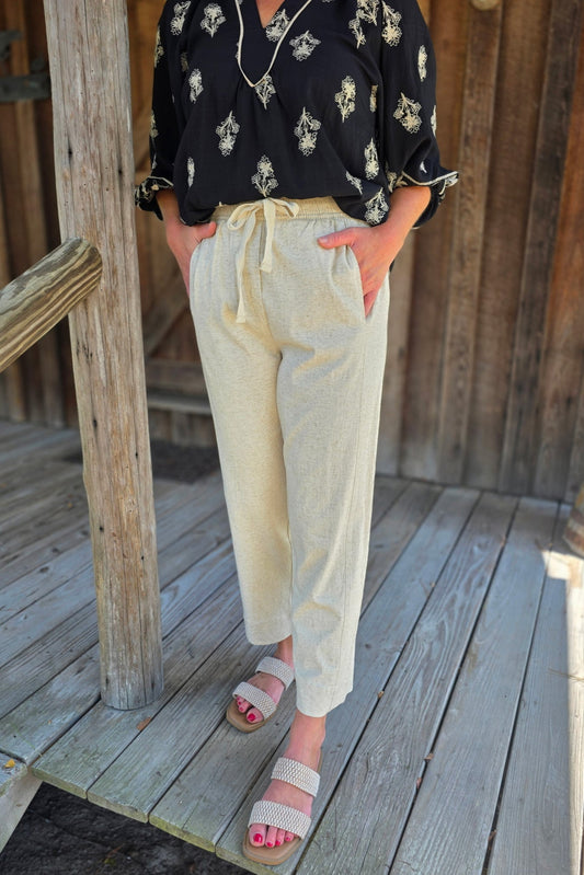 Woman wearing light beige linen pants with a drawstring waist and side pockets, styled with a black printed blouse and woven slide sandals.