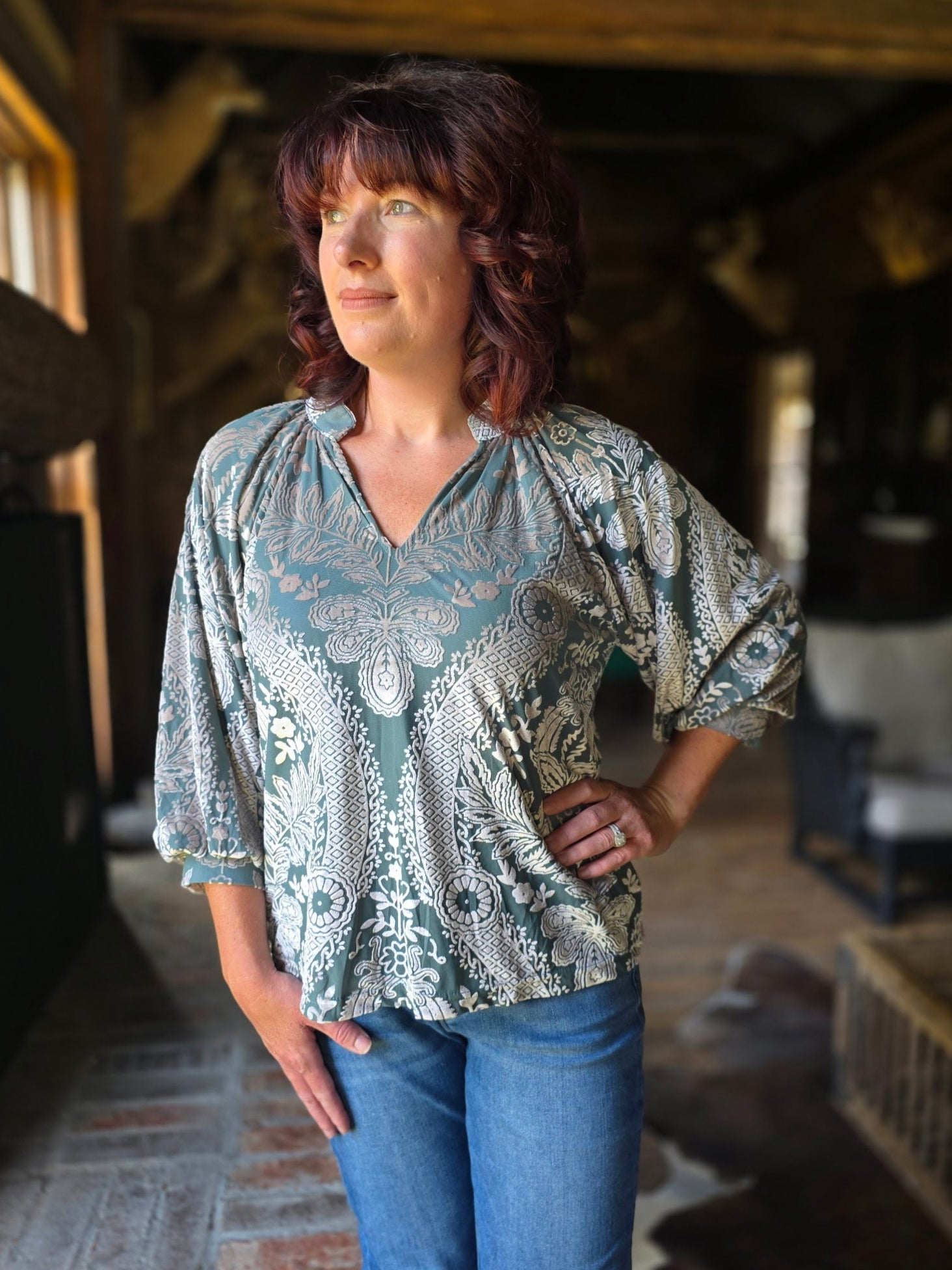 Woman wearing a patterned blouse and blue jeans standing in a rustic interior setting.