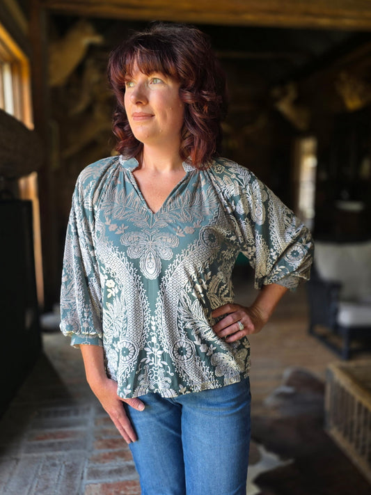 Woman wearing a patterned blouse and blue jeans standing in a rustic interior setting.