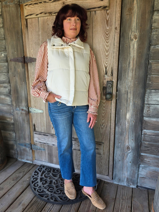 Woman wearing an ivory zip-up puffer vest styled with a striped blouse and denim jeans, standing on a rustic porch.