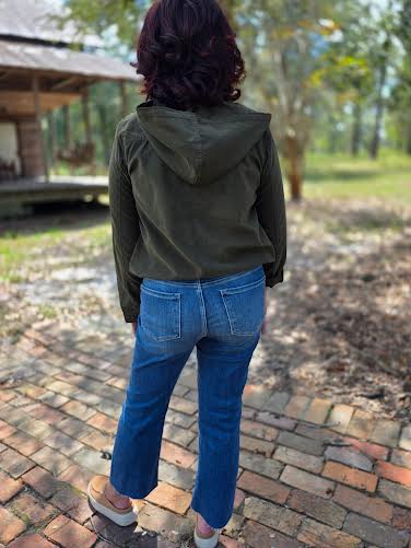 Person wearing a green jacket and blue jeans standing on a brick path with trees in the background