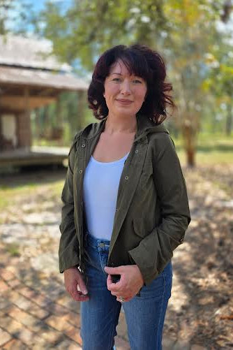 Woman standing outdoors in a natural setting wearing an olive green jacket.